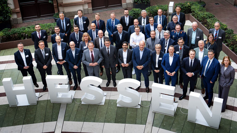 Über 30 Personen stehen in 3 Reihen nebeneinander in einem Innenhof und schauen nach oben in die Kamera. Vor ihnen sind 1 Meter hohe Buchstaben platziert, die das Wort "Hessen" ergeben.