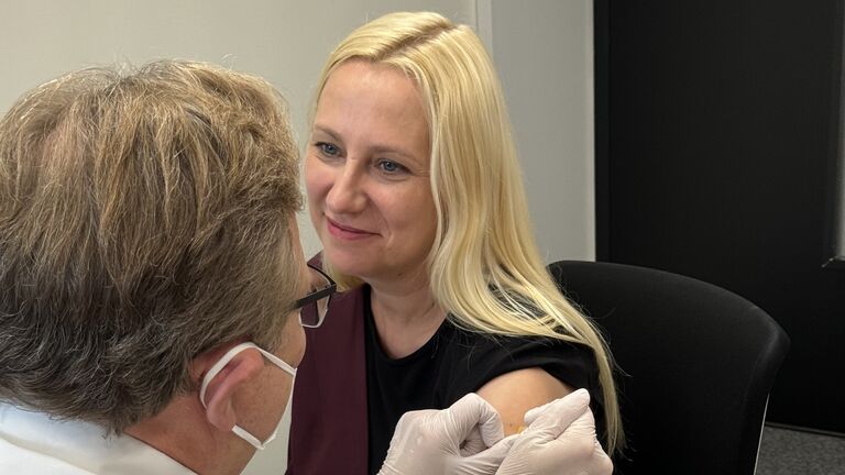 Gesundheitsministerin Stolz sitzt auf einem Stuhl. Ein Arzt ist gerade dabei ein Pflaster auf ihren Oberarm zu kleben.