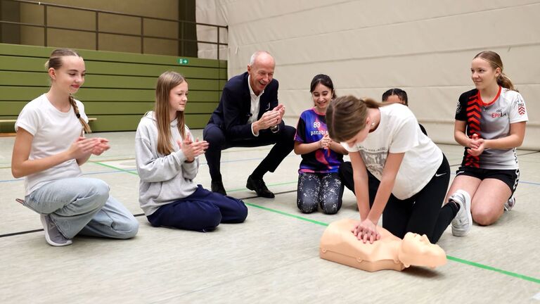 Mehrere Schülerinnen knien in Turnhalle. Eine Schülerin übt an Puppe Wiederbelebung.