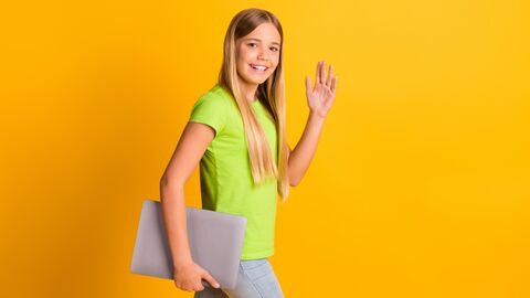 Mädchen lauft an einer gelben Wand entlang. Sie trägt ein Laptop unter dem Arm und winkt in die Kamera
