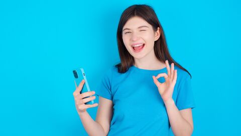 Mädchen in blauem Shirt steht vor einer blauen Wand. Sie zwinkert, hält ein Handy in der einen Hand und macht ein TOP Zeichen mit der anderen