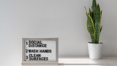Symbolbild Corona-Regeln Ein Schild mit der Aufschrift "1 Social Distance 2 Wash Hands 3 Clean Surfaces" steht auf einem hellgrauen Sideboard. Daneben steht eine Topfpflanze in einem weißen Umtopf.
