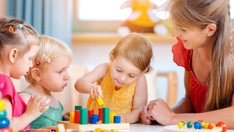 Eine Erzieherin spielt mit drei Kindern am Tisch mit bunten Klötzchen.