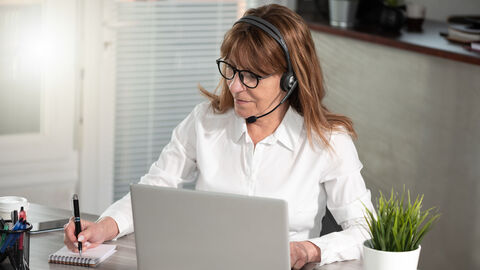 Eine Frau mit Headset sitzt vor einem Laptop und schreibt auf einem Notizblock.
