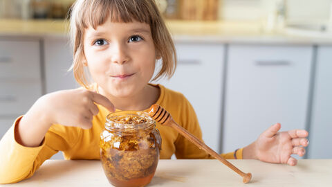 Hmmmm, leckerer Honig! Ein Kind sitzt am Küchentisch und lässt sich Honig aus dem Glas schmecken.