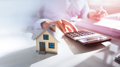 Ein kleines Holzhäuschen im Vordergrund, dahinter eine Person am Schreibtisch mit Taschenrechner und Unterlagen vor sich