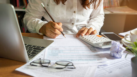 Finanzen Eine Frau sitzt an einem Schreibtisch, vor ihr ein Laptop, Unterlagen und ein Taschenrechner.