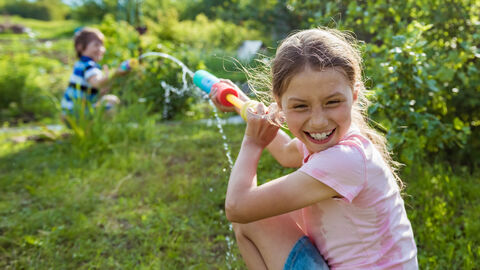 So macht der Sommer Spaß! Zwei lachende Kinder bespritzen sich auf eienr Wiese mit Wasser.