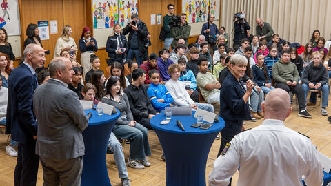 In einem Raum sitzen viele Schüler aufgereiht zusammen, vor ihnen stehen moderierende Personen und Innenminister Poseck sowie Kultusminister Schwarz.