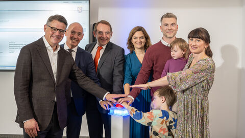 V. l. n. r.: Ministerpräsident Boris Rhein, WIBank-Sprecher Gottfried Milde, Finanzminister Alexander Lorz, WIBank-Geschäftsführerin Judith Mandel und die Hessengeld-Empfänger-Familie, Tobias und Isabel Zöller mit ihren Kindern Sechs Erwachsene und zwei Kinder stehen in einem Halbkreis und strecken jeweils einen Arm in die Mitte.