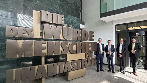 Justizminister Heinz steht mit drei weiteren Männern neben dem Schriftzug zur Menschenwürde