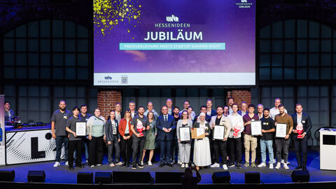 Mehrere Personen stehen nebeneinander auf einer Bühne. Im Hintergrund wird auf eine Leinwand "Hessen Ideen Jubiläum" projiziert.