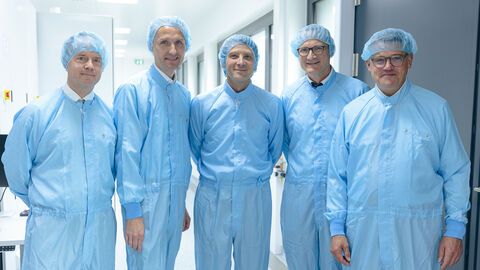 Fünf Personen stehen in blauen Schutzanzügen nebeneinander in einem hellen Raum.