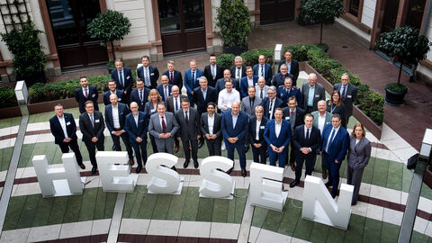 Über 30 Personen stehen in 3 Reihen nebeneinander in einem Innenhof und schauen nach oben in die Kamera. Vor ihnen sind 1 Meter hohe Buchstaben platziert, die das Wort "Hessen" ergeben.