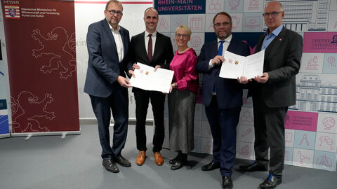 v.l.n.r.: Prof. Dr. Enrico Schleiff (Präsident der Goethe-Universität Frankfurt und amtierender RMU-Sprecher), Clemens Hoch (Rheinland-Pfälzischer Minister für Wissenschaft und Gesundheit), Prof. Dr. Tanja Brühl (Präsidentin der TU Darmstadt), Timon Gremmels (Hessischer Minister für Wissenschaft und Forschung, Kunst und Kultur) und Prof. Dr. Georg Krausch (Präsident der Johannes Gutenberg-Universität Mainz).  Fünf Personen stehen nebeneinander und halten zwei Urkunden hoch.