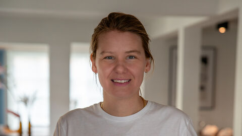 Eine Frau trägt ein weißes Oberteil und lächelt in die Kamera. Der Hintergrund ist verschwommen.