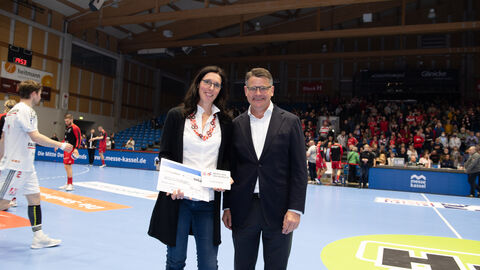 Ministerpräsident Boris Rhein überreicht Carolin Goujard für ihren ehrenamtlichen Einsatz die Auszeichnung "Menschen des Respekts". Ministerpräsident Boris Rhein steht auf dem Spielfeld der MT Melsungen. Die Ehrenamtliche Carolin Goujard hat von ihm eine Auszeichnung überreicht bekommen. Diese hält sie in der Hand.