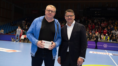 Ministerpräsident Boris Rhein überreicht Uwe Lohmann für seinen ehrenamtlichen Einsatz die Auszeichnung "Menschen des Respekts". Ministerpräsident Boris Rhein steht auf dem Spielfeld der MT Melsungen. Der Ehrenamtliche Uwe Lohmannhat von ihm eine Auszeichnung überreicht bekommen. Diese hält er in der Hand.