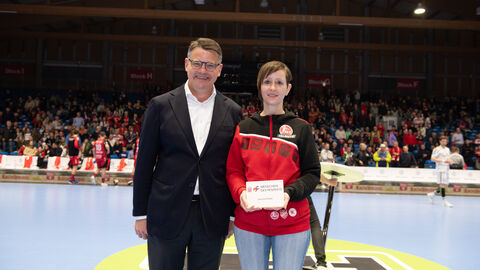 Ministerpräsident Boris Rhein überreicht Jessica Fetzer für ihren ehrenamtlichen Einsatz die Auszeichnung "Menschen des Respekts". Ministerpräsident Boris Rhein steht auf dem Spielfeld der MT Melsungen. Die Ehrenamtliche Jessica Fetzer hat von ihm eine Auszeichnung überreicht bekommen. Diese hält sie in der Hand.