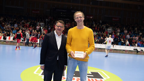 Ministerpräsident Boris Rhein überreicht Stefan Döring für seinen ehrenamtlichen Einsatz die Auszeichnung "Menschen des Respekts". Ministerpräsident Boris Rhein steht auf dem Spielfeld der MT Melsungen. Der Ehrenamtliche Stefan Döring hat von ihm eine Auszeichnung überreicht bekommen. Diese hält er in der Hand.
