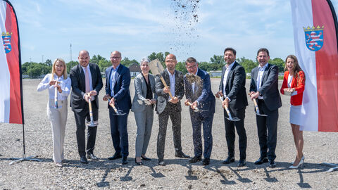 Ministerspräsident Rhein steht mit weiteren Personen zwischen zwei Hessen-Flaggen auf einem Baugrundstück. Alle halten Schaufeln in Händen und werfen teilweise Erde in die Luft.