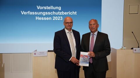 Vorstellung Verfassungsschutzbericht Hessen 2023 Innenminister Poseck hält Vefassungsschutzbericht in Kamera. Im Hintergrund steht geschrieben: Vorstellung Verfassungsschutzbericht Hessen 2023