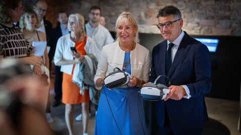 Digitalministerin Sinemus und Ministerpräsident Rhein mit VR-Brille Digitalministerin Sinemus und Ministerpräsident Rhein halten VR-Brillen in Kamera. Im Hintergrund sieht man weitere Menschen.
