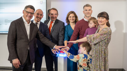 V. l. n. r.: Ministerpräsident Boris Rhein, WIBank-Sprecher Gottfried Milde, Finanzminister Alexander Lorz, WIBank-Geschäftsführerin Judith Mandel und die Hessengeld-Empfänger-Familie, Tobias und Isabel Zöller mit ihren Kindern. Sechs Erwachsende und zwei Kinder stehen in einem Halbkreis und strecken jeweils einen Arm in die Mitte.