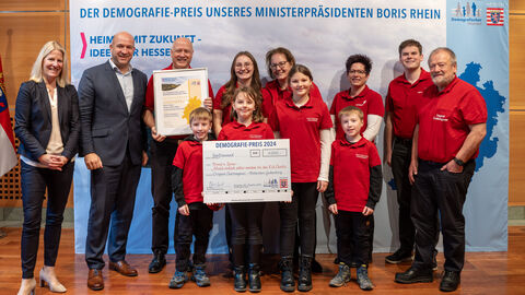 2. Preis: Original Chattengauer-Bläserchor Gudensberg – Brass‘n’Spass Musik einfach selber machen bei den Kid.Chatts Eine Gruppe Personen steht vor einem Aufsteller mit der Aufschrift "Der Demografie-Preis unseres Ministerpräsidenten Boris Rhein". Eine Person hält eine gerahmte Urkunde in Händen, eine weitere einen Scheck.