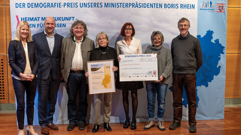 3. Preis: Kunst in Licher Scheunen e.V., Lich – Kunst in Licher Scheunen Eine Gruppe Personen steht vor einem Aufsteller mit der Aufschrift "Der Demografie-Preis unseres Ministerpräsidenten Boris Rhein". Eine Person hält eine gerahmte Urkunde in Händen, eine weitere einen Scheck.