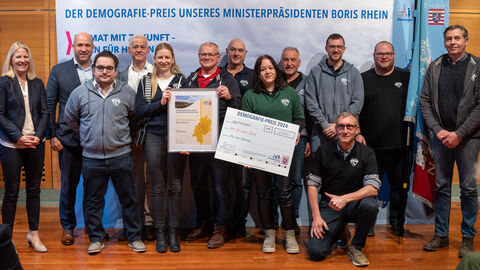 3. Preis: „Mir sein Seldersch“, Selters im Taunus – Wir für unser Dorf Eine Gruppe Personen steht vor einem Aufsteller mit der Aufschrift "Der Demografie-Preis unseres Ministerpräsidenten Boris Rhein". Zwei Personen halten eine gerahmte Urkunde in Händen, eine weitere einen Scheck.