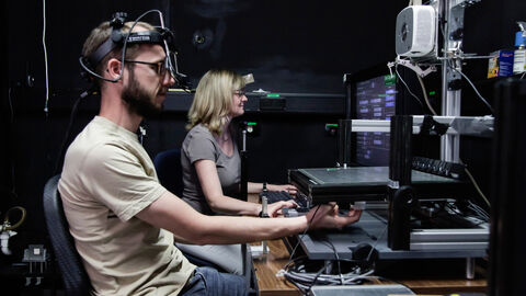 Zwei Personen in einer Laborsituation: Der Proband trägt eine Konstruktion auf dem Kopf, die ihn mit dem PC vor ihm verbindet, die Forscherin sitzt neben ihm.