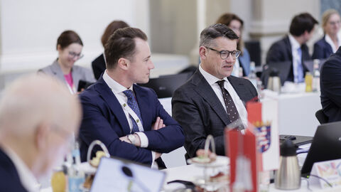 Zwei Männer sitzen in Anzügen nebeneinander. Der rechte schaut nach vorne, während der linke den anderen ansieht.