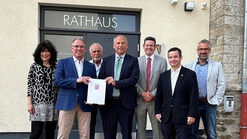 Eine Frau und mehrere Männer stehen vor einem Gebäude mit Aufschrift "Rathaus" und lächeln in Kamera. Zwei der Männer halten einen Zuwendungsbescheid in den Händen.