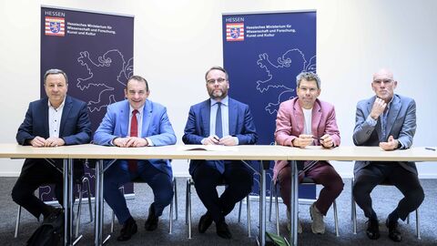 Sie sehen von links nach rechts: Prof. Dr. Karim Khakzar (Sprecher der Hessischen Hochschulen für Angewandte Wissenschaften),  Prof. Dr. R. Alexander Lorz (Hessischer Finanzminister), Timon Gremmels (Hessischer Minister für Wissenschaft und Forschung, Kunst und Kultur), Prof. Dr. Thomas Nauss (Sprecher der Konferenz Hessischer Universitätspräsidien), Prof. Elmar Fulda (Sprecher der Hessischen Kunsthochschulen) Fünf Personen sitzen an einem Tisch, im Hintergrund Plakate mit dem Wappen des Hessischen Ministeriums für Wissenschaft und Forschung, Kunst und Kultur